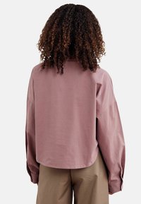 Pārmērīgs, gaiši mauve krekls ar apaļu apakšmalu, nolaistām plecu daļām un garām piedurknēm. Gluda kokvilnas struktūra; vienkāršs dizains.