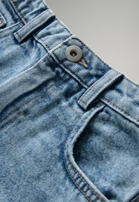 Világoskék farmernadrág texturált felülettel. Jellemzői a fém gomb, az övbújtatók és a derékrész és zsebek mentén látható varrás.