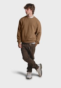 Brauner Sweatshirt mit Rundhalsausschnitt und gerippten Bündchen, kombiniert mit dunkelbraunen Hosen und grauen Turnschuhen. Das Model posiert in entspannter Haltung.