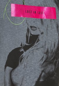 Grijze T-shirt met een zwart-witafbeelding van het gezicht van een vrouw en een roze "VERLOREN IN LUST" lint, omrand door groene cirkelvormige lijnen.