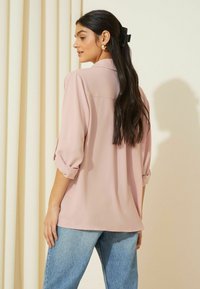 Helles rosa Bluse aus glattem Stoff, mit lockerer Passform und hochgerollten Ärmeln. Kombiniert mit mittelblauen Jeans.