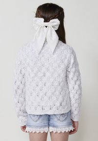 Pull en tricot blanc avec motif ajouré, associé à un short en denim avec broderie en dentelle. Cheveux attachés avec un grand nœud en satin blanc.