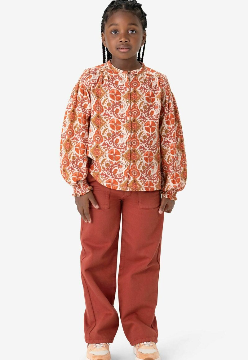 Blouse à manches longues à fleurs en orange et crème, avec un devant à boutons, associée à un pantalon ample couleur rouille et des baskets légères.