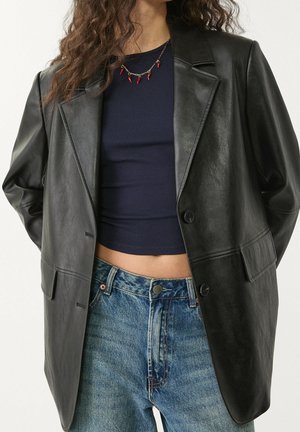 Vrouw draagt een oversized zwarte leren blazer over een cropped navy top en high waist blauwe jeans, met een ketting van rode chilipeper.