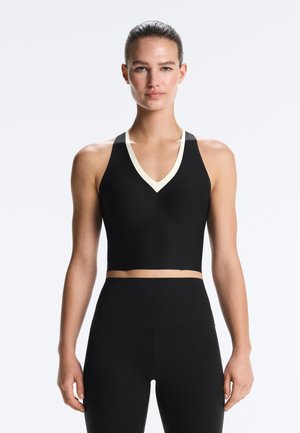 Kobieta ubrana w czarny, bezrękawnikowy krótki top z białym dekoltem w kształcie litery V oraz czarne legginsy z wysokim stanem, stojąca na tle jednolitego, jasnego tła.