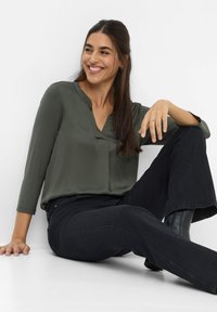 Blusa verde oliva a maniche lunghe con scollatura a V e una leggera lucentezza, abbinata a jeans neri a campana e stivali neri alla caviglia. Tessuto con una texture liscia.