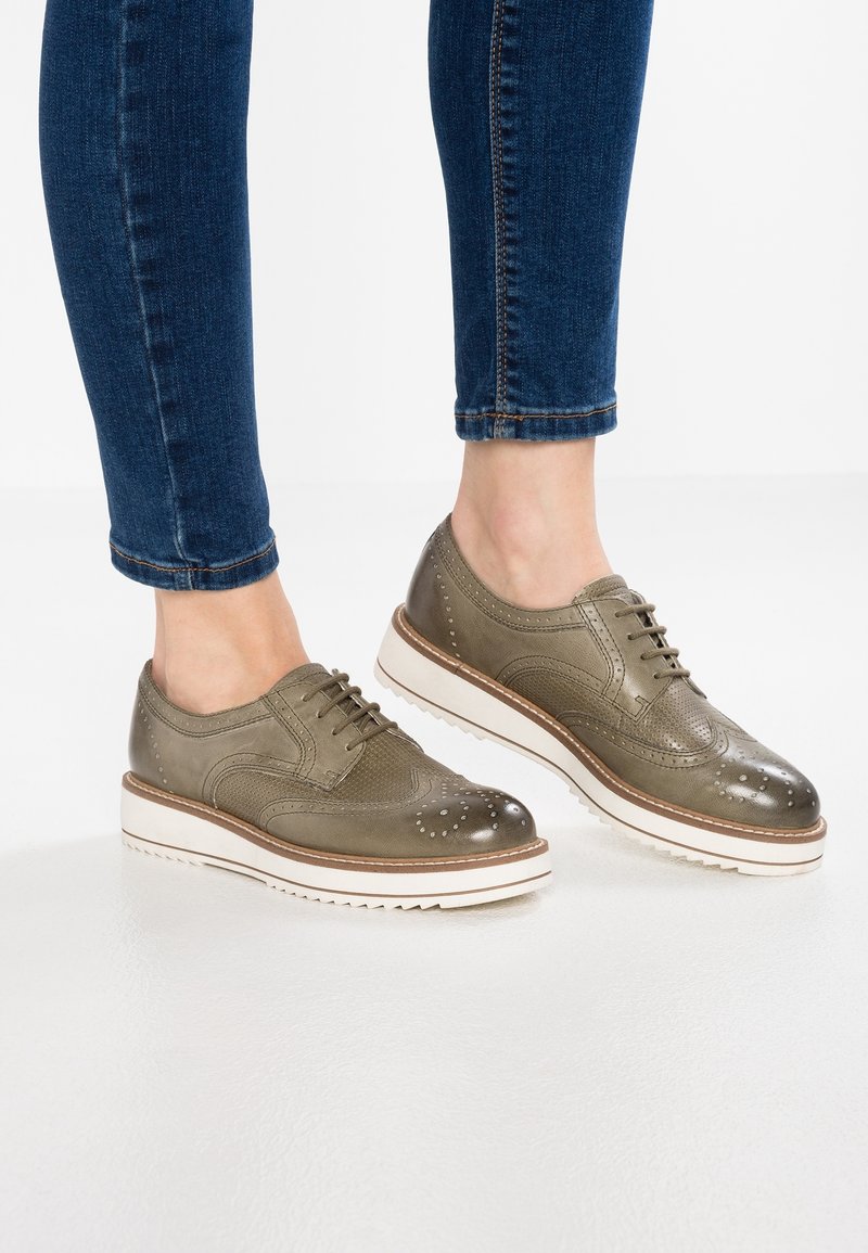 Femme portant des chaussures brogues en cuir vert olive avec des semelles compensées blanches et un jean skinny bleu sur fond blanc.