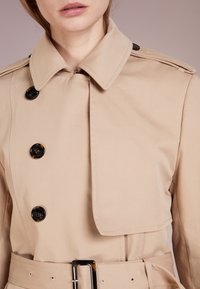 Cappotto trench beige con colletto, chiusura doppiopetto, bottoni neri e vita con cintura. Tessuto liscio e vestibilità su misura con spalline.