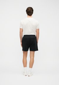 Μαύρο σορτς με πίσω τσέπη, σε συνδυασμό με λευκή μπλούζα T-shirt. Το μοντέλο φαίνεται από πίσω, παρουσιάζοντας μια άνετη εφαρμογή και ομαλή υφή υφάσματος.