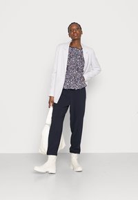 Blazer branco, blusa com padrão floral em azul marinho, calças pretas e botas brancas até o tornozelo. Modelo segura uma bolsa branca. Design simples e clean.