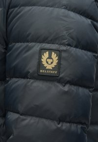 Laivastonsininen toppatakki, jossa on tikattu kuvio. Rintakehässä kullanvärinen merkki, jossa lukee "BELSTAFF". Sileä pinta.