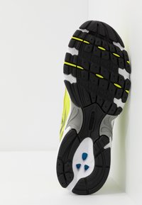 Svart och grå atletisk skosula med ett texturerat mönster, med ljus gula detaljer och en vit stödkonstruktion under.