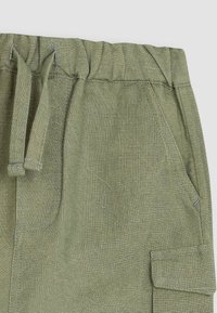 Zelené lněné kalhoty s elastickým pasem, detaily na zavazování a bočními kapsami. Textura se zdá být lehká a prodyšná.