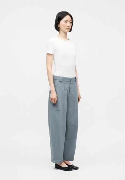 Γυναίκα που στέκεται φορώντας λευκό τοπ με κοντά μανίκια, φαρδιά γκρι παντελόνια με ψηλή μέση και μεγάλες τσέπες, και μαύρα επίπεδα παπούτσια σε απλό φόντο.