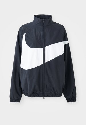 Nike vindjakke i mørkeblå med en fremtredende hvit swoosh. Har høy krage, glidelås foran og elastiske mansjetter. Lett, strukturert stoff.