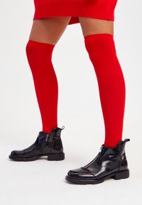 Botas de tobillo de charol negro con cremallera frontal y acentos texturizados, combinadas con vibrantes medias rojas hasta el muslo. Superficie suave, diseño elegante.