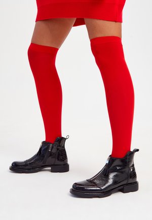 Black patent ankle boots with a front zipper and textured accents, paired with vibrant red thigh-high socks. Smooth surface, sleek design.