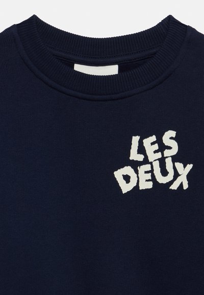 Тъмносиня блуза с кръгло деколте с бял текстурален надпис "LES DEUX" на горната част на гърдите.