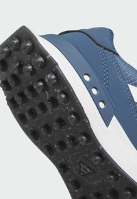 Niebieski but sportowy z teksturowaną cholewką, białymi akcentami oraz czarną gumową podeszwą z licznymi chwytającymi kolcami zapewniającymi przyczepność.