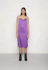 Uma mulher em pé, vestindo um vestido midi roxo sem mangas com alças finas e botas pretas até o tornozelo, contra um fundo claro.