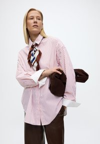 Γυναίκα με μακριά ξανθά μαλλιά που φοράει χαλαρό πουκάμισο με κόκκινες ρίγες, μεταξωτό κασκόλ με σχέδια και καφέ δερμάτινο παντελόνι, κρατώντας ένα διπλωμένο καφέ σακάκι.