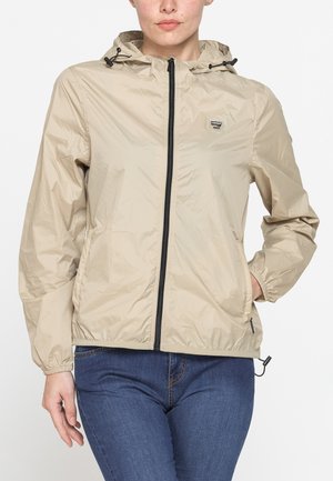 Donna che indossa una giacca beige con cappuccio e zip, polsini elastici e jeans blu su sfondo bianco uniforme.
