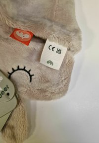 Esquina de un suave muñeco de peluche beige con bordado de ojos cerrados, una etiqueta roja con un icono de hipopótamo y una etiqueta blanca que muestra las marcas CE y UKCA.