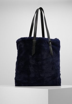 Marineblå taske i imiteret pels med to lange sorte skulderremme i læder og spændedetaljer, vist på en hvid piedestal.