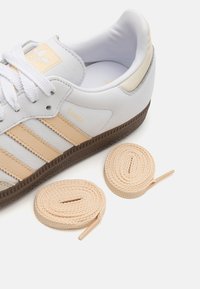 Fehér bőrsneaker világos bézs részletekkel, három csíkkal, egy bordó gumitalppal, és mellette két bézs cipőfűzővel.