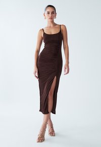 Robe midi marron sans manches avec fronces sur le côté et une fente haute sur la cuisse. Texture lisse, coupe cintrée, associée à des talons beiges à brides.