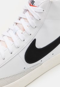 Vit sneaker i mjukt läder, med en svart mocka Nike-swoosh, vita snören, en texturerad grå gummisula och stickade detaljer.