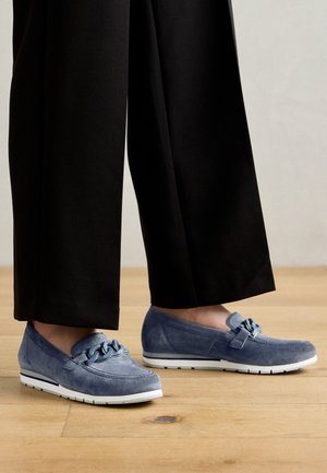 Pieds chaussés de mocassins en daim bleu avec des semelles épaisses blanches et des chaînes décoratives, assortis à un pantalon noir à jambes larges sur un sol en bois.