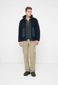 Mlad čovjek u mornarskoplavoj flis jakni, maslinastozelenoj zip-up majici, bež cargo hlačama i crnim tenisicama, stoji protiv obične bijele pozadine.