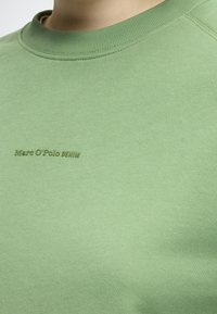 Sweatshirt de algodão verde-claro com decote canelado. Logótipo "Marc O'Polo DENIM" bordado num verde ligeiramente mais escuro. Textura suave.