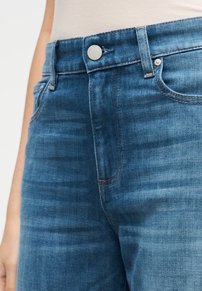 Jeans di denim blu con chiusura frontale a bottoni, cinque tasche e cuciture a contrasto arancioni sulle cuciture. Tessuto liscio con sfumature sottili.