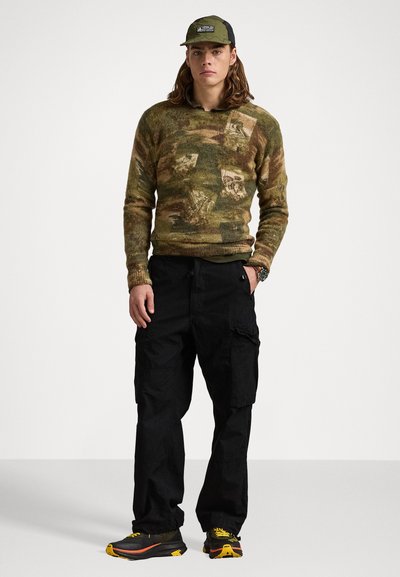 Groen patroon gebreide trui, zwarte cargobroek en veelkleurige sneakers. De outfit heeft zakken en een pet met een logo.