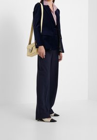 Blazer in velluto blu navy, camicia rosa con colletto, pantaloni formali a gamba larga blu navy, décolleté a punta crema e nere, e una borsa a tracolla beige.