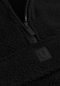 Čierny fleecový pulóver s textúrovanou tkaninou, polovičným zipsom a obdĺžnikovou čiernou nášivkou na hrudi. Hladký detail zipsu.