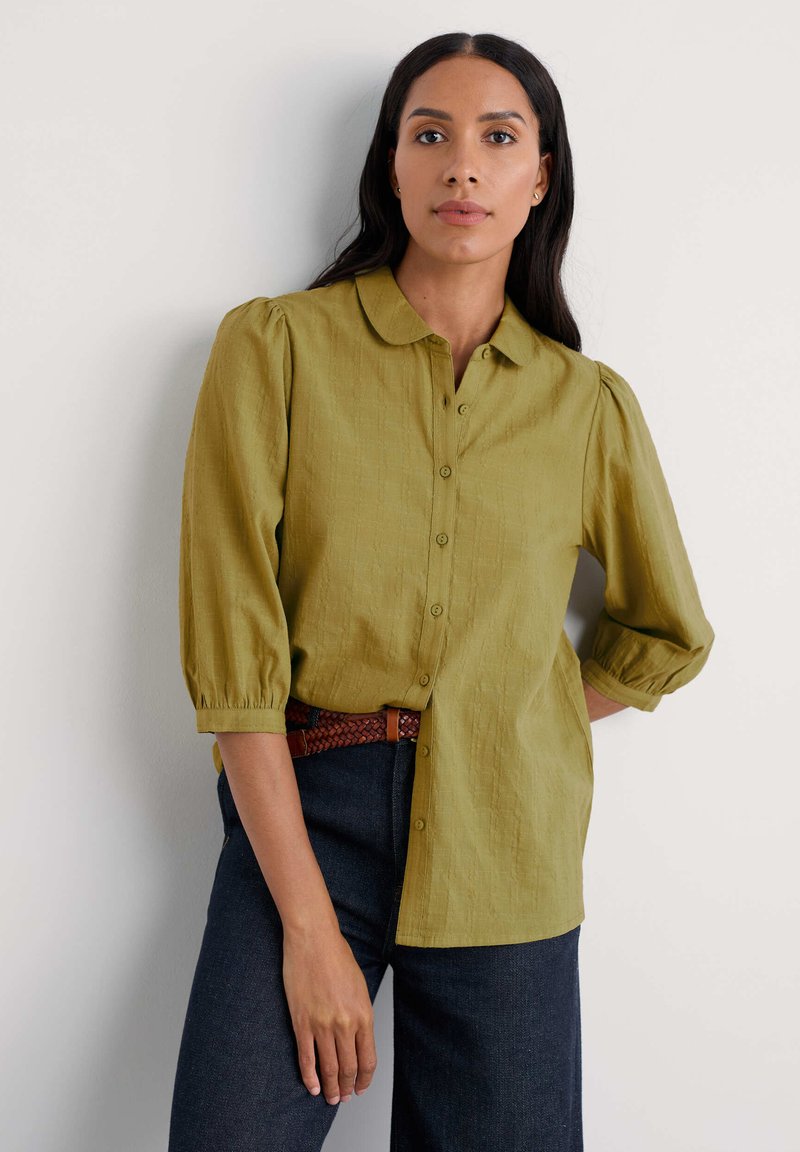 Oliwkowo-zielona bluzka na guziki z kołnierzykiem, bufiastymi rękawami i fakturowaną tkaniną. Połączona z ciemnymi dżinsami i plecionym brązowym paskiem.