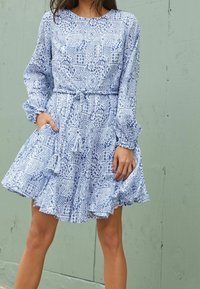 Robe bleu clair avec un motif, manches longues, ceinture à la taille et ourlet à volants. Le tissu semble doux avec une silhouette fluide.