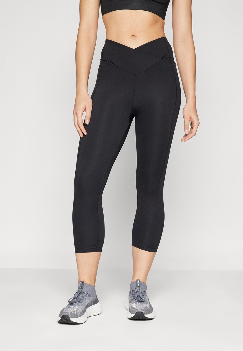 I leggins neri a capri presentano un design con vita incrociata. Il tessuto ha una superficie liscia ed è abbinato a scarpe da ginnastica grigie.