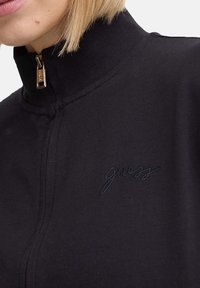 Chaqueta negra con cremallera, que cuenta con un alto cuello, cremallera oculta y el logotipo bordado "guess" en hilo a juego en el pecho. Textura suave.