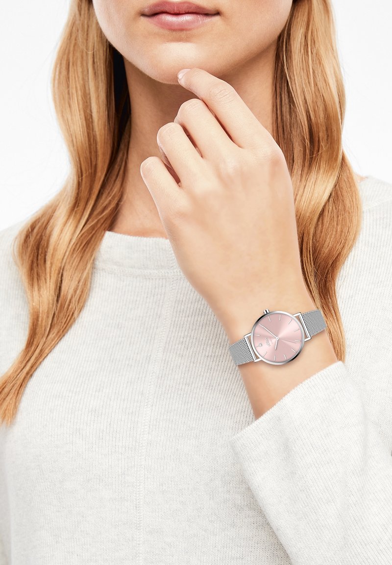 Femme aux longs cheveux blonds portant un pull clair, montrant une montre argentée avec un cadran rose à son poignet près de son visage.
