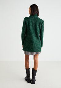 Manteau vert texturé avec un col montant, des manches longues et une silhouette ajustée, superposé sur une jupe grise et des bottes noires.