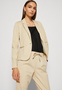 Blazer beige ajusté avec un col cranté, un bouton, et deux poches frontales, associé à un haut noir ample et un pantalon beige assorti.