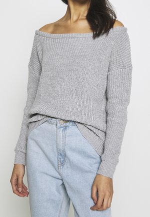 Nainen, joka pukeutuu harmaaseen olkapäät paljastavaan neulepaitaan, joka on työnnetty vaaleansinisiin korkeavyötäröisiin farkkuihin, seisoo yksivärisen taustan edessä.