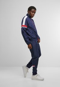 Navy tracksuit dengan garis putih dan merah di lengan serta bagian bawah celana, terbuat dari kain halus. Model mengenakan sepatu sneaker putih.