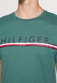 Camiseta de algodón verde que presenta un gran logo en azul marino de "HILFIGER" y una franja horizontal en rojo, blanco y azul marino a lo largo del pecho.