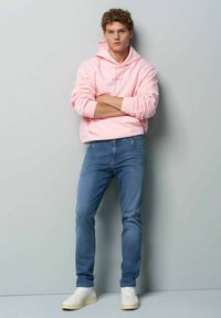 Lichtroze hoodie met een trekkoord, gecombineerd met blauwe jeans en witte sneakers. De hoodie heeft ribgebreide manchetten en een ontspannen pasvorm.