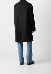Abrigo negro largo con un diseño a medida, que presenta un cuello alto y puños con botones, llevado sobre unos vaqueros azul claro y zapatos negros.